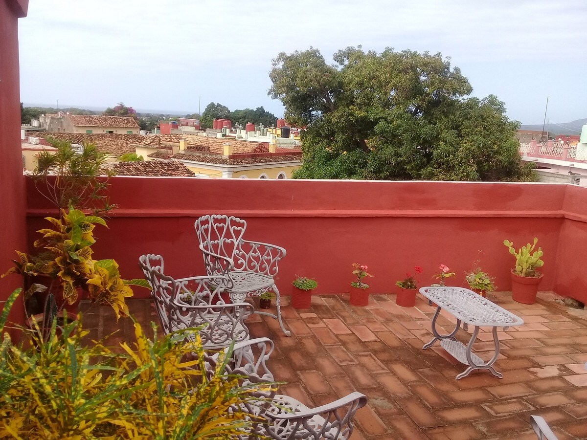 A rooftop terrace features decorative metal seating around a small table. Potted plants and colorful flowers adorn the area, while a view of the cityscape with terracotta rooftops and distant greenery is visible in the background.