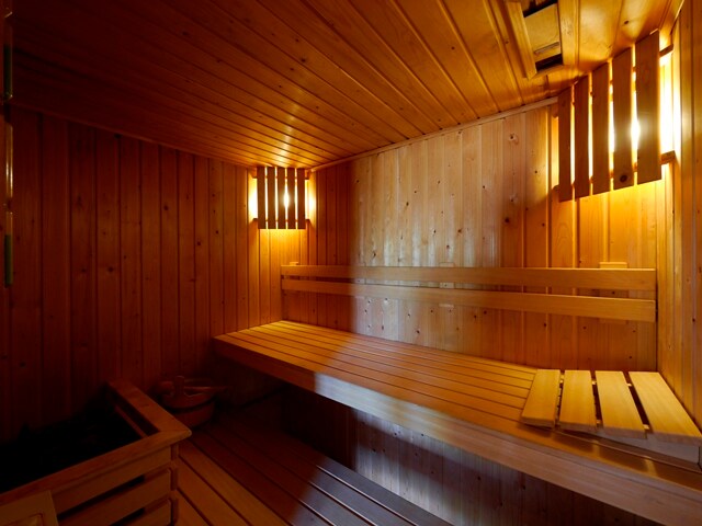 A wooden sauna interior features warm, soft lighting and smooth wooden benches. The walls and ceiling are paneled with light wood, creating a cozy atmosphere. A small area for hot stones is visible, enhancing the tranquil and relaxing environment.