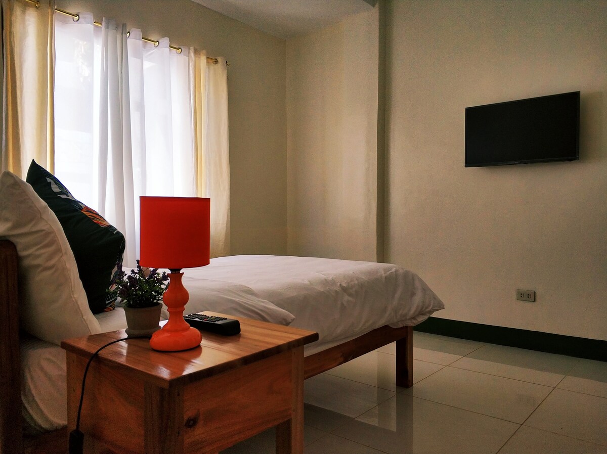 A bright sleeping area features a spacious bed dressed in crisp white linens. Natural light is provided by large windows with sheer curtains. A wooden bedside table holds a vibrant orange lamp and a small decorative plant, while a flat-screen TV is mounted on the wall.