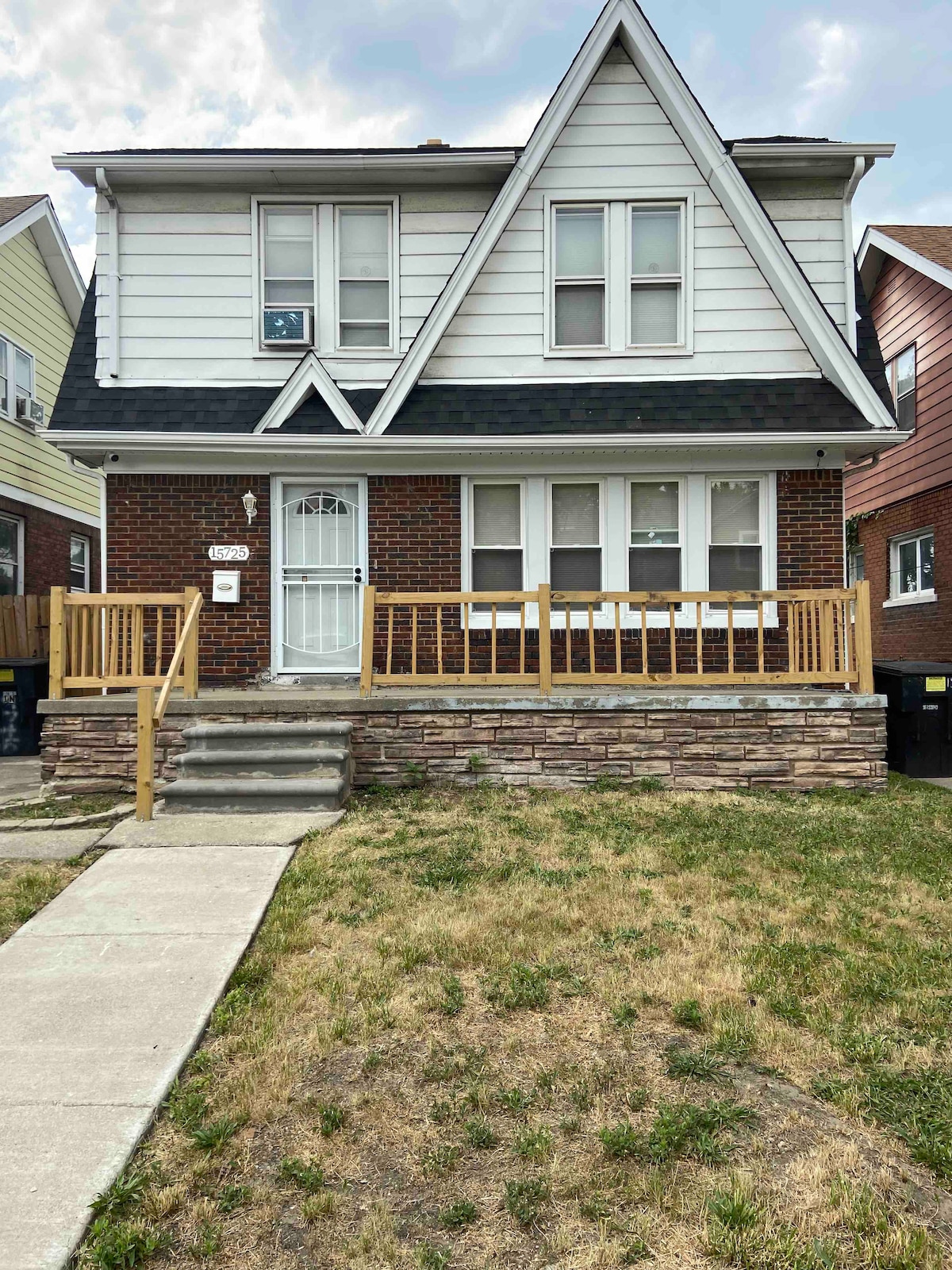 A two-story house with a mix of brick and siding exterior is presented. The front porch features a railing and steps leading to the main entrance. The yard is mostly grassy, with a concrete walkway leading up to the house, surrounded by neighboring homes.