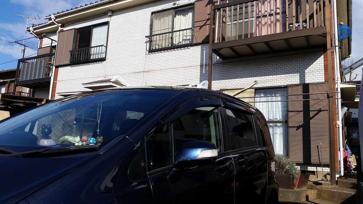 A two-story apartment building is featured with distinct balconies on the upper level. A parked black vehicle is positioned in the foreground, providing convenient access. Windows allow natural light to brighten the rooms, while a decorative touch is added by plants near the entrance.