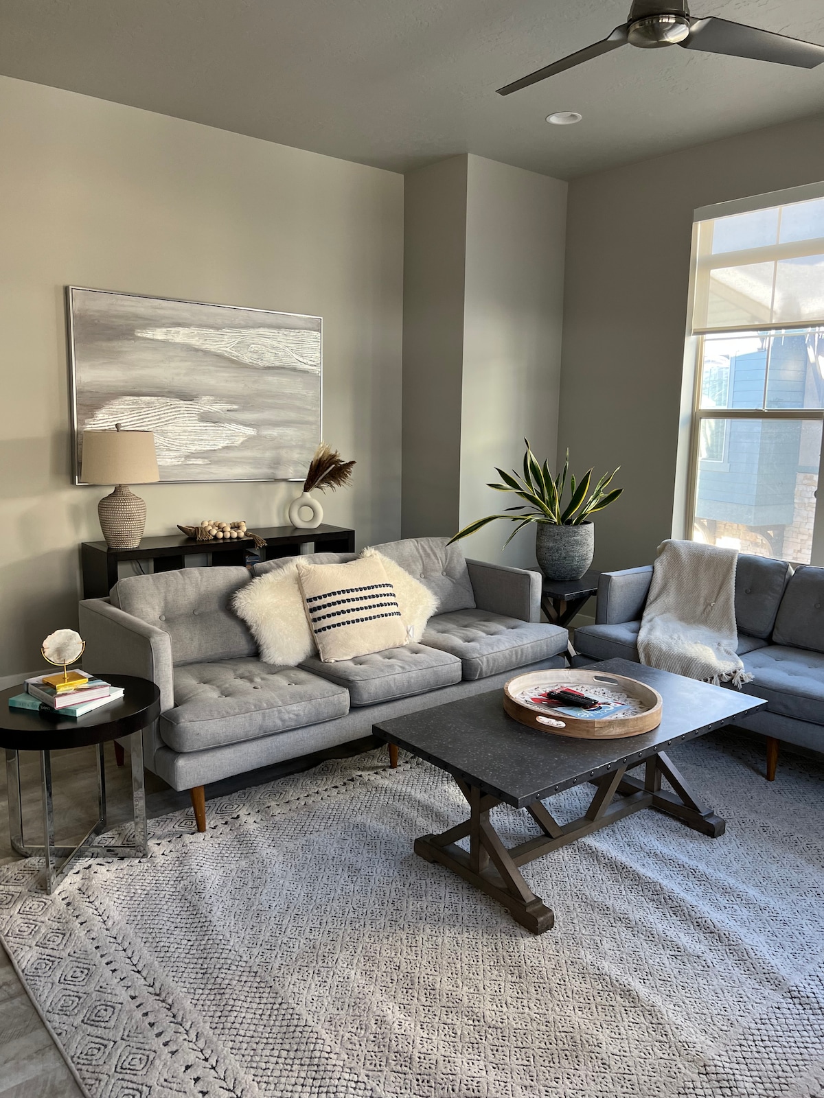 The living area features a grey sectional sofa complemented by a coordinating armchair. A dark wood coffee table sits on a light woven rug, with decorative items arranged atop it. A tall potted plant stands beside the sofa, with natural light entering through large windows.