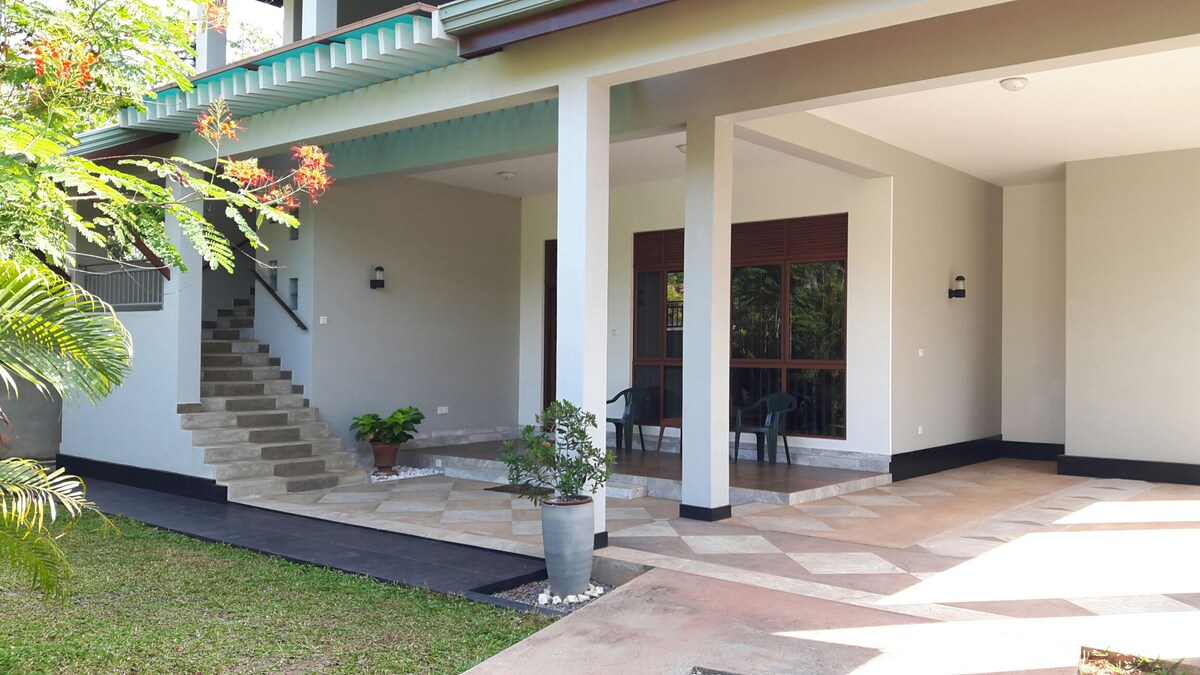 A spacious outdoor area features a covered patio with seating arrangements and a potted plant. The entrance leads to a landscaped garden, while stone steps provide access to the upper levels. Natural light illuminates the entryway, enhancing the tranquil setting.