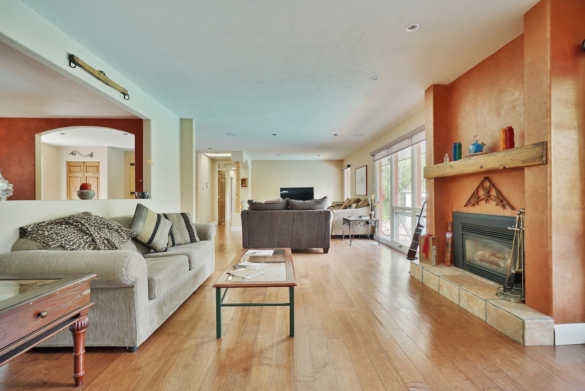 A spacious living area features light wood flooring and a cozy fireplace. The seating arrangement includes two comfortable sofas and a coffee table. Large glass doors lead to an outdoor area, while soft natural light illuminates the room, enhancing its open layout.