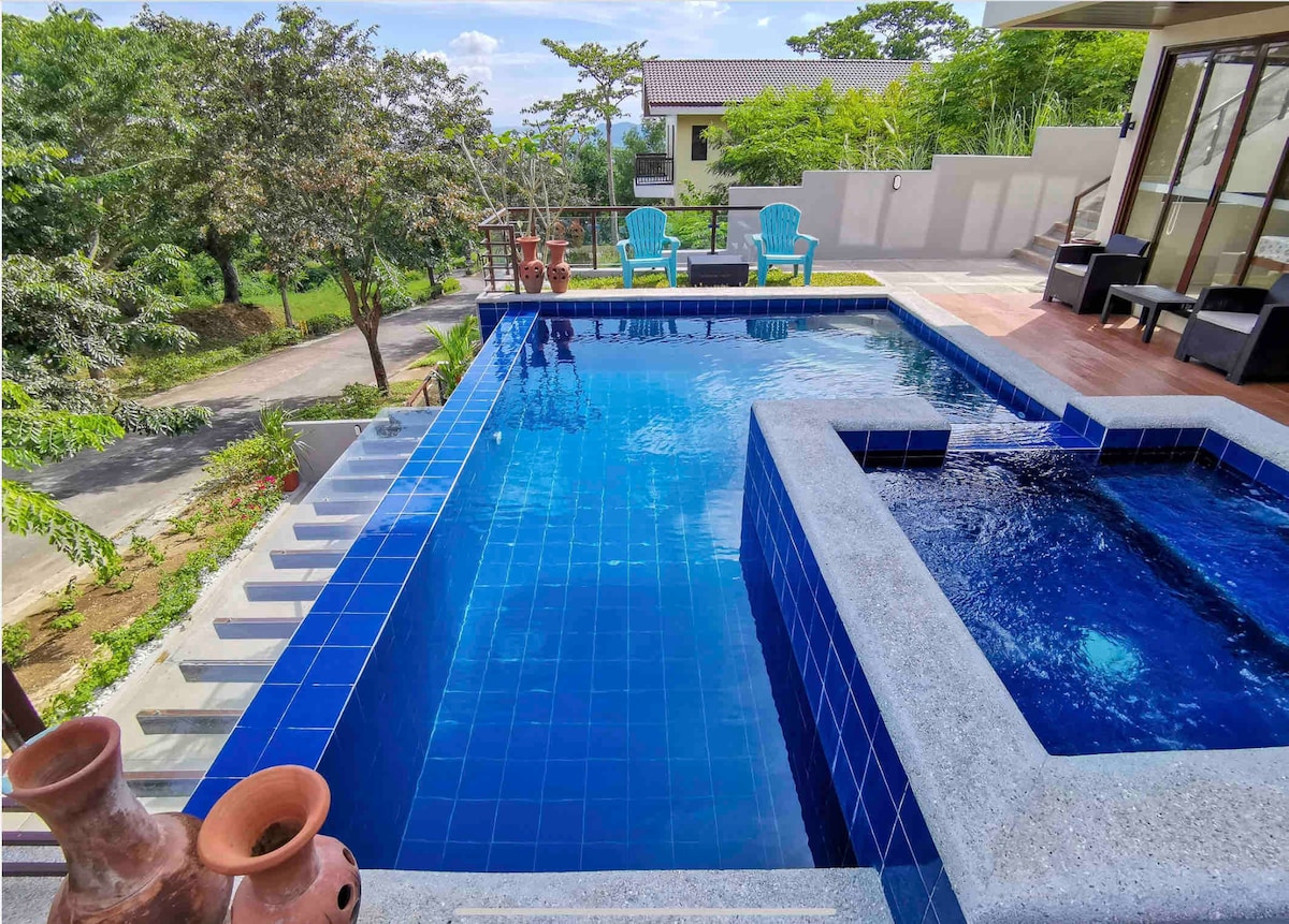 An outdoor infinity pool is complemented by a heated hot tub, surrounded by a landscaped area. Two blue lounge chairs are positioned nearby, inviting relaxation. The pool's deep blue tiles reflect the sky, while steps lead down from the pool area. The surroundings feature greenery and a tranquil view.