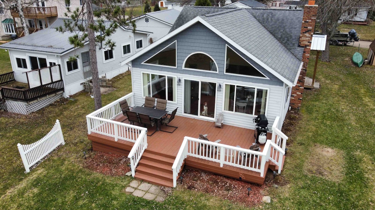 An expansive deck surrounds the cottage, featuring a dining table and lounge chairs for outdoor relaxation. Large windows provide views of the surroundings, while a well-maintained lawn is visible. The cottage’s architecture includes a prominent arched window, enhancing the inviting outdoor space.