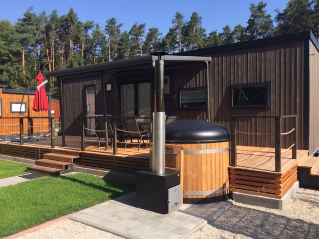 The exterior of the mobile home features a spacious wooden terrace with seating arrangements and a red shade umbrella. A Finnish bathing tub is positioned alongside the terrace, complemented by a stylish metal chimney and surrounding greenery.
