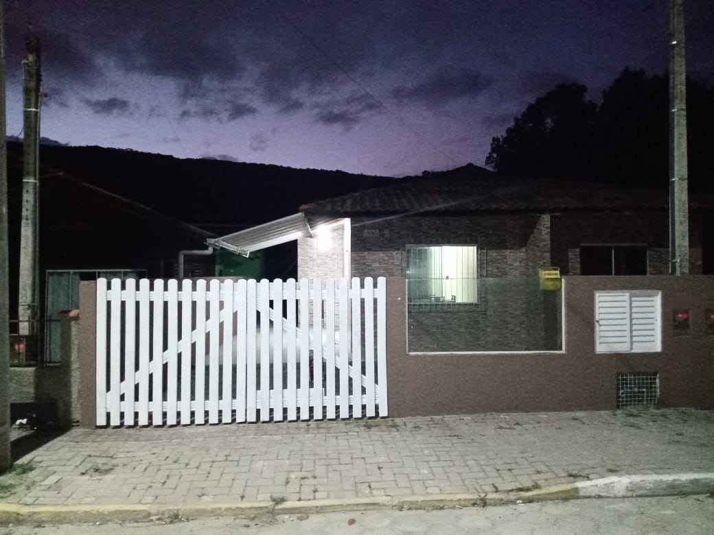The exterior of a single-story house is displayed at dusk, featuring a white picket fence and a lighted entrance. The brick facade is complemented by large windows, providing a view of the interior. Surrounding scenery includes trees and a paved street.