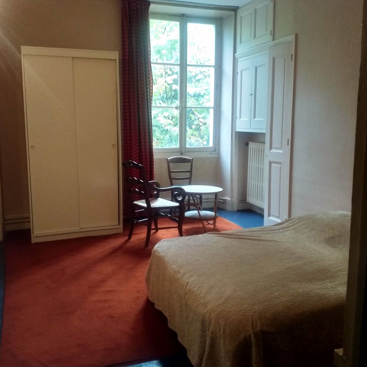 A spacious room features a large bed covered with a light blanket. A small table and a chair occupy the center, while a wardrobe stands against one wall. Natural light enters through a window dressed with simple curtains, illuminating the room's neutral tones.