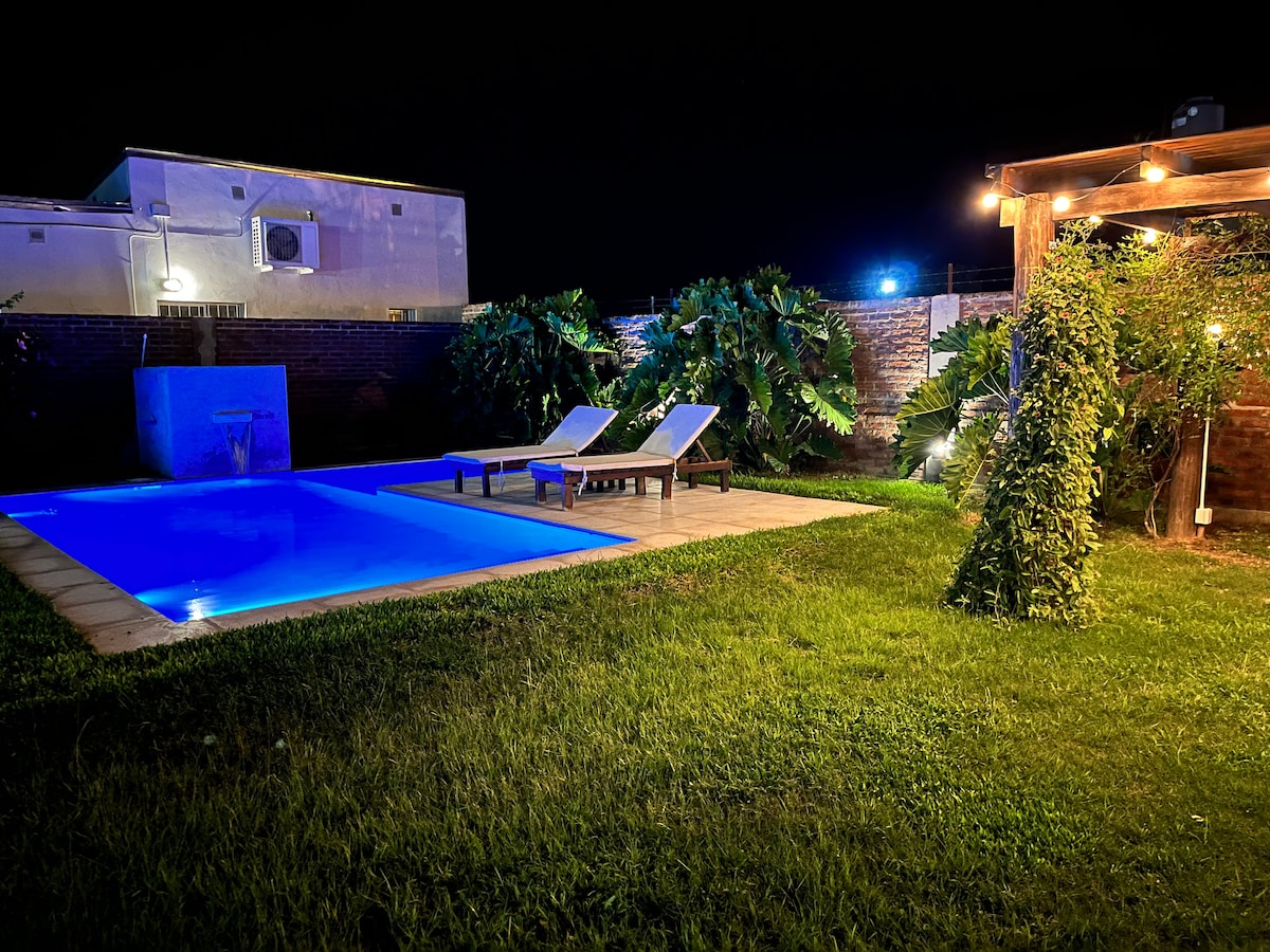 A well-lit outdoor area features a tranquil pool surrounded by lush greenery. Two lounge chairs are positioned beside the water, with a soft glow emanating from nearby lights. A charming waterfall adds a calming element to the scene.
