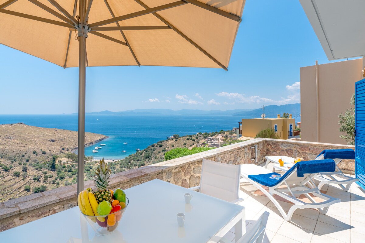 An outdoor terrace features a shaded dining table with a bowl of fresh fruit, overlooking the sparkling blue waters of Klima Bay. Sun loungers are arranged nearby, with a scenic view of the coastline and boats dotting the horizon under a clear blue sky.