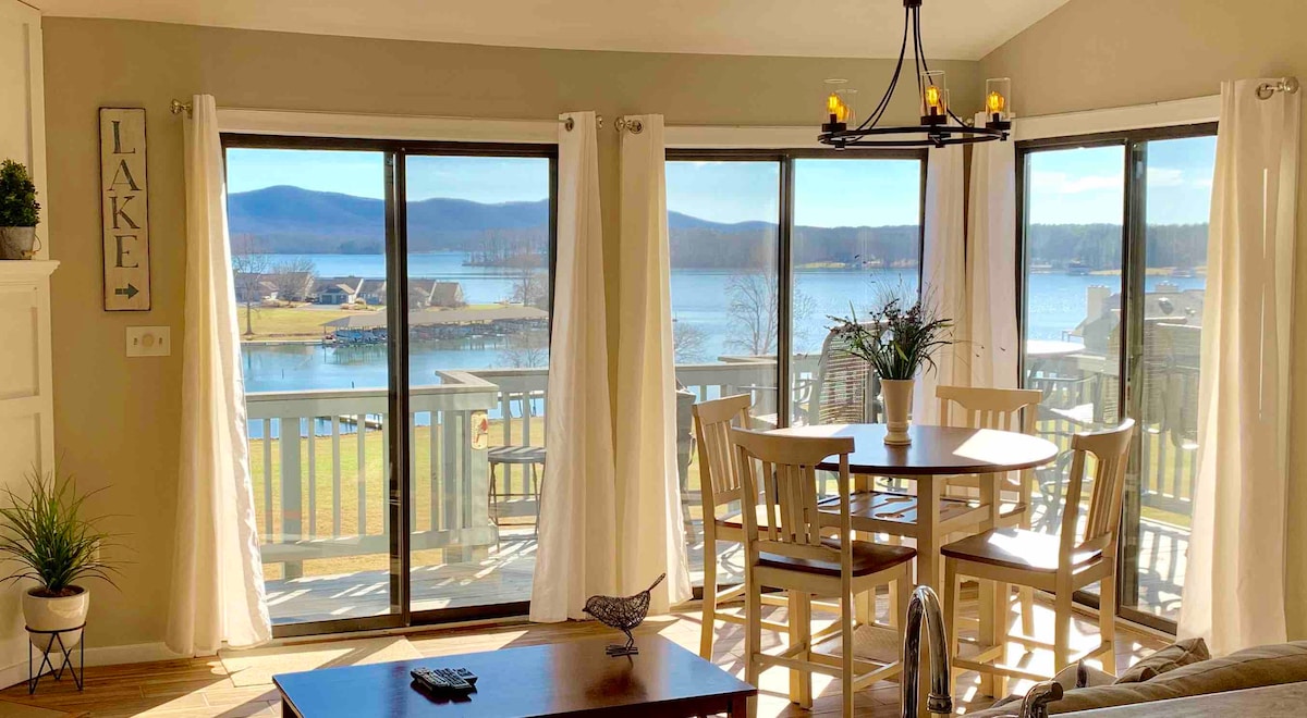 A dining area features a round table surrounded by four light-colored chairs. Large sliding glass doors provide unobstructed views of the lake and surrounding mountains, while natural light fills the space through sheer white curtains. Decorative items include a plant and a simple centerpiece.