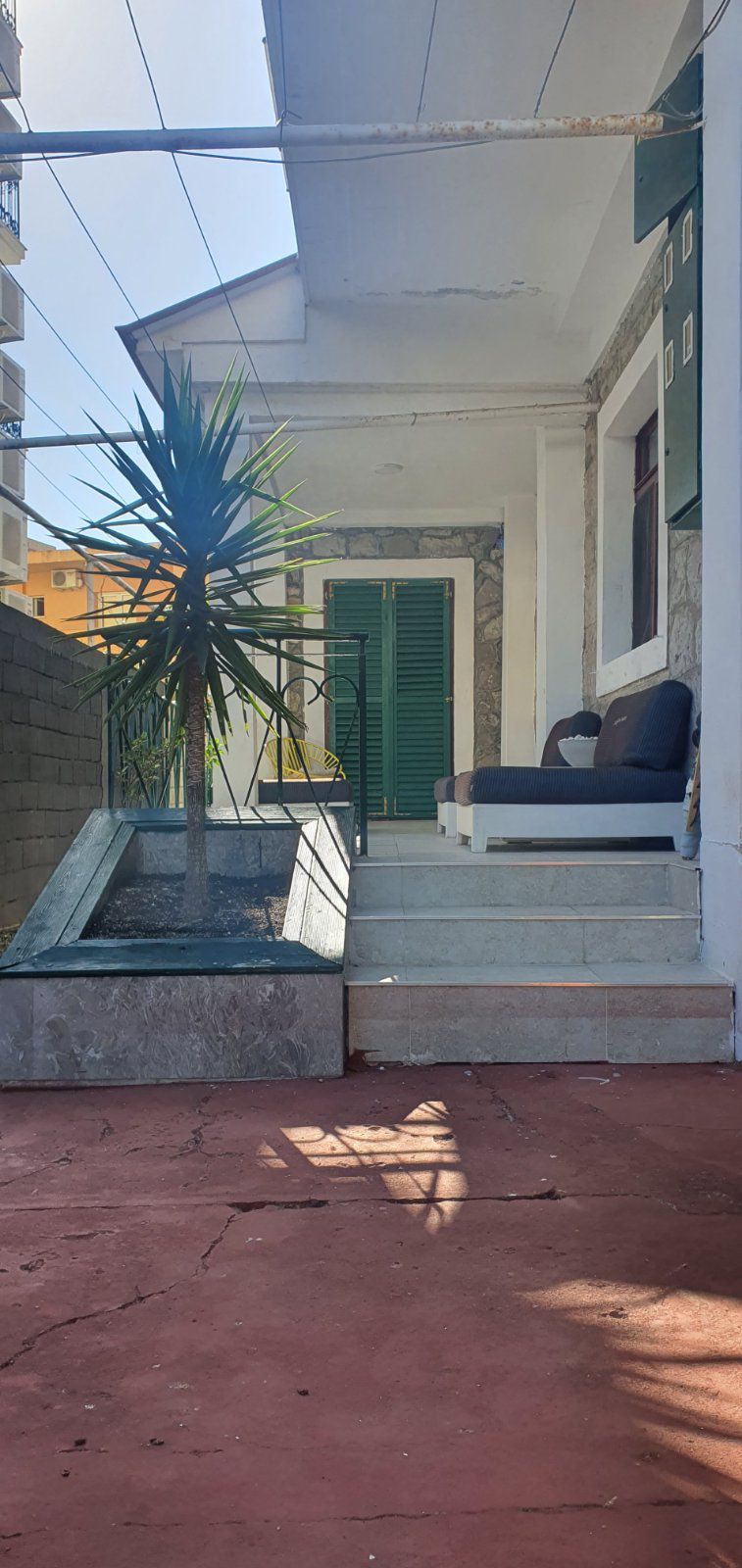 A patio area is visible, featuring a simple seating arrangement next to a small potted plant. Sunlight casts shadows on the red floor, while green shutters adorn the entrance, adding a touch of color to the space.