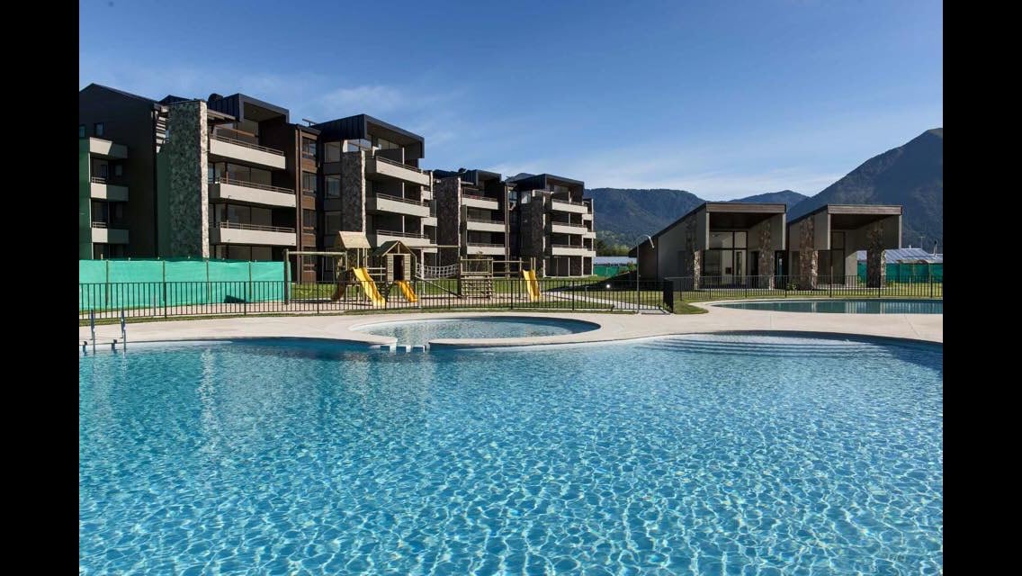 A clear view of three buildings within the Costa Pucón condominium, surrounded by a vibrant blue pool. The pool area features play structures, and the distant mountains serve as a scenic backdrop under a bright sky.