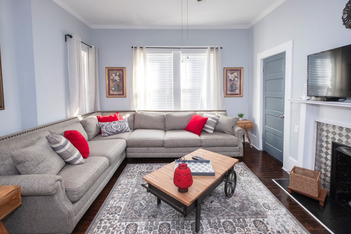 A spacious living room features a large gray sectional sofa adorned with red accent pillows. Natural light streams through the window treatments, illuminating a wooden coffee table with a decorative red lantern. Framed artworks and a decorative fireplace add character to the inviting space.