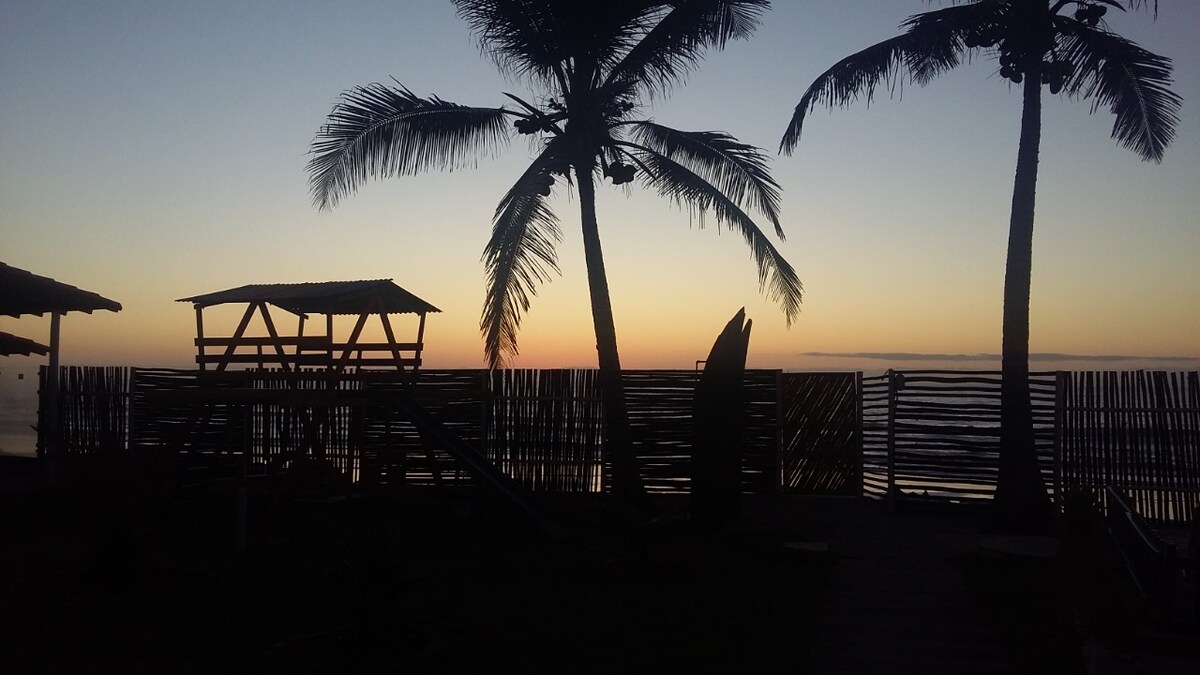 Silhouetted palm trees frame a serene beach scene at dusk. A wooden play structure is visible in the foreground, with a rustic fence and calm water in the background, reflecting soft hues of sunset.