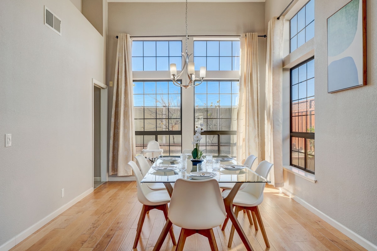 A spacious dining area features a modern glass table surrounded by six chairs. Large windows allow natural light to flood the room, accentuated by tall curtains. A minimalistic chandelier hangs above the table, creating an inviting space for meals.