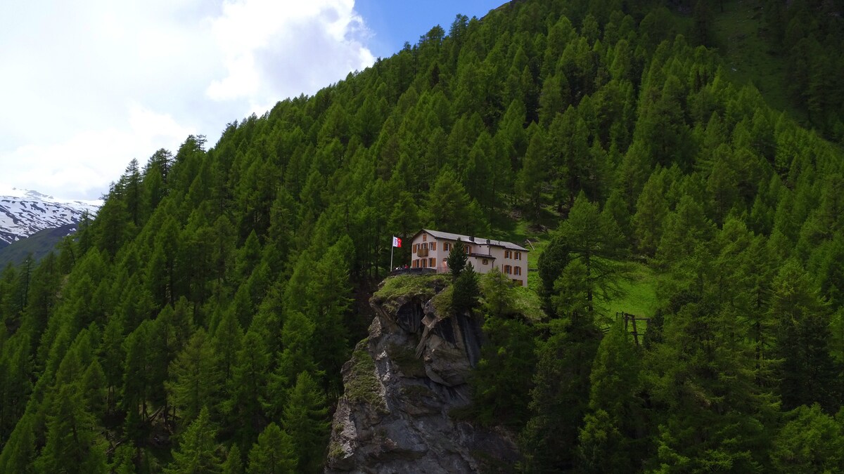 The mountain hotel is perched on a rocky outcrop, surrounded by vibrant green trees and mountainous terrain. A flag is visible, and the natural beauty of the location is highlighted by the distant snow-capped peaks under a partly cloudy sky.