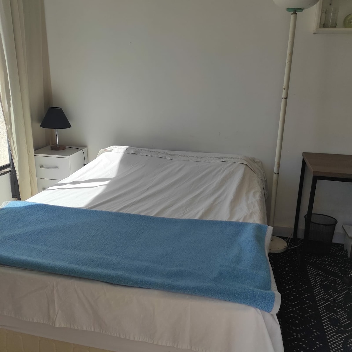 A well-lit sleeping area features a spacious bed with a light-colored blanket draped over it. A calming blue towel is placed at the foot of the bed. A bedside lamp and a small white nightstand are positioned beside the bed, complementing the minimalist decor.