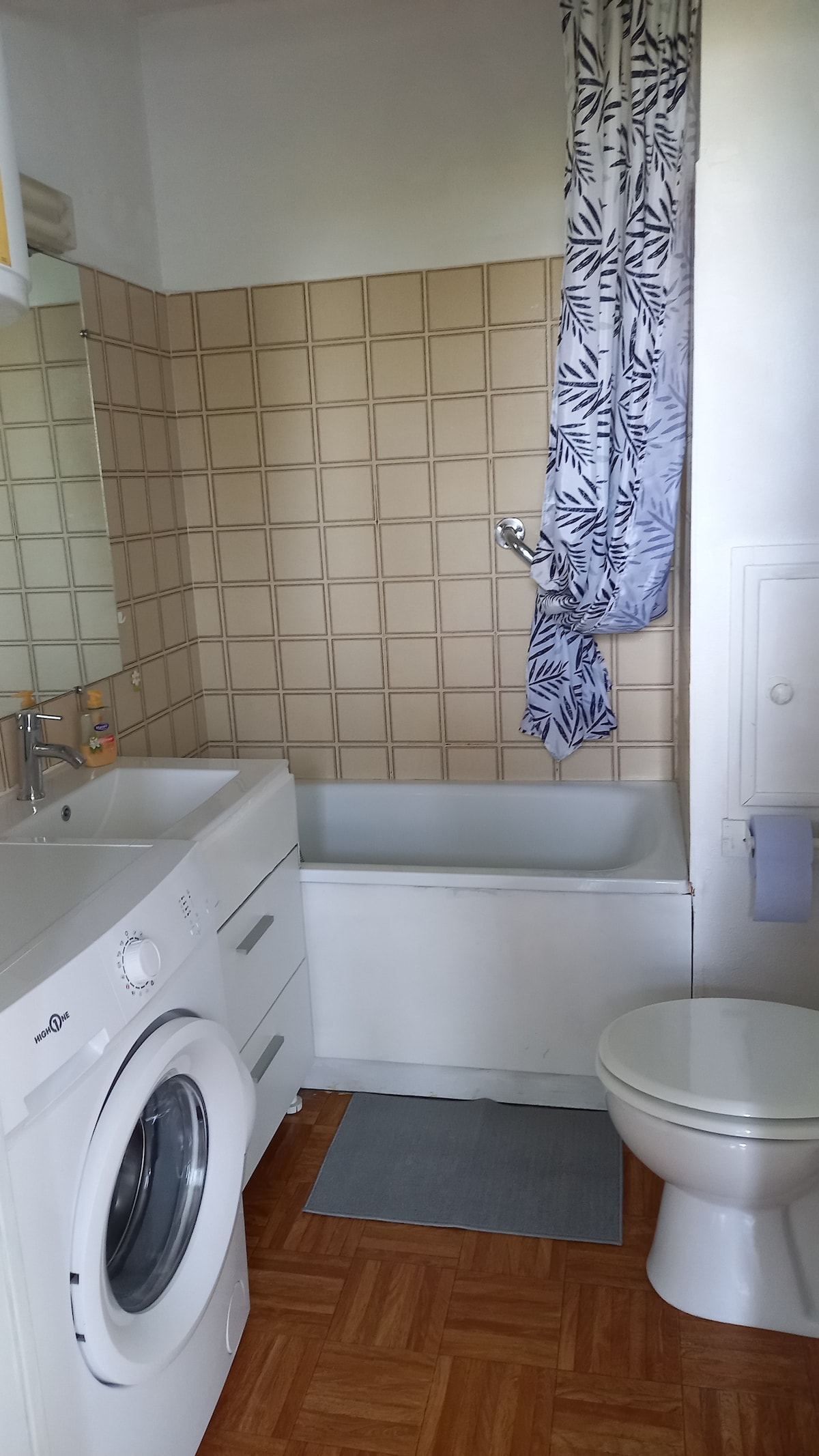 The bathroom features a bathtub with a showerhead above, complemented by patterned curtains. A washing machine is integrated within the cabinetry, and a toilet is present. The flooring is a honey-colored wood, while tiles with a neutral tone cover the walls.
