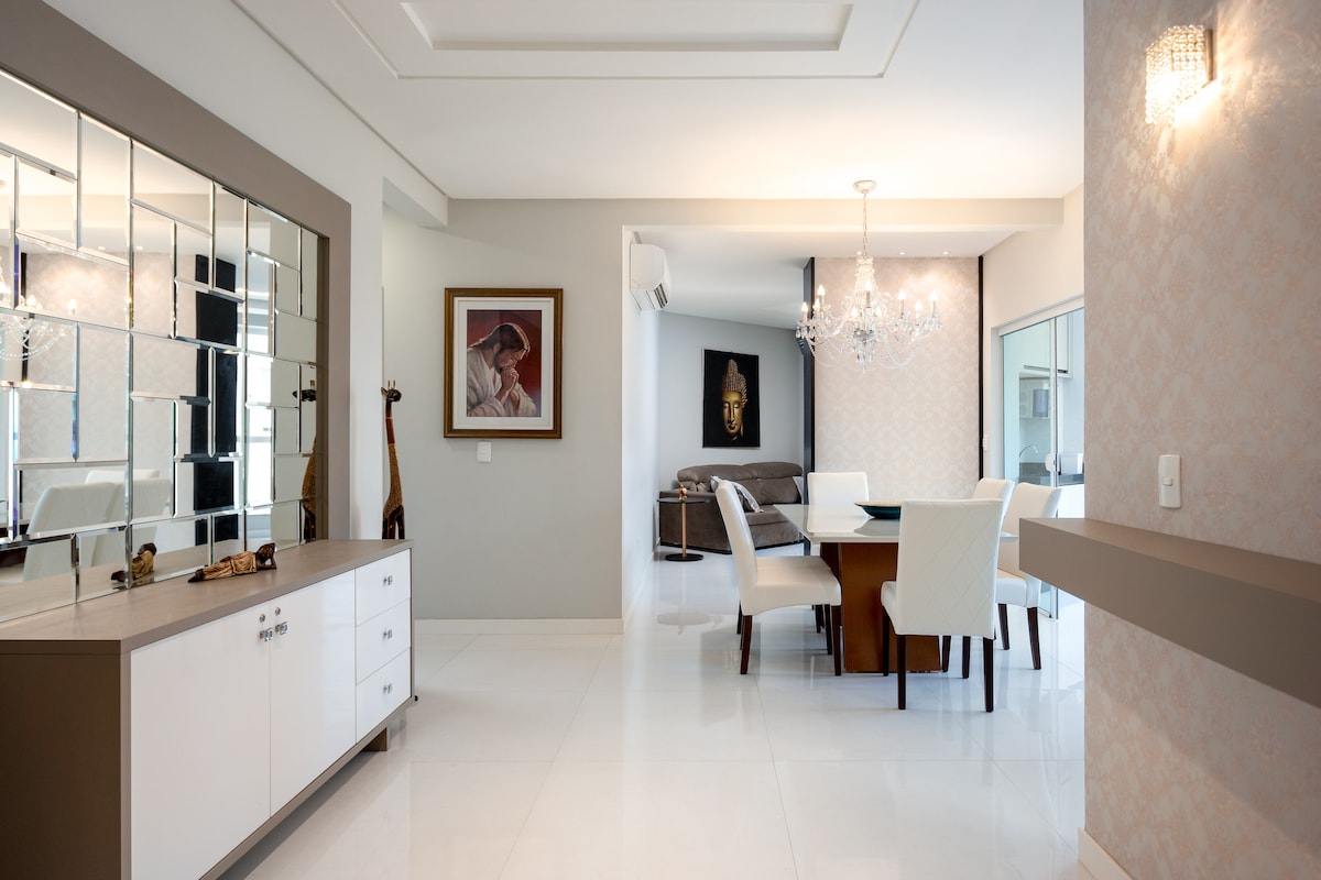 A modern interior showcases an open-plan dining and living area. A sleek white sideboard displays decorative items, while a dining table is set for six. Natural light brightens the space, highlighting the elegant light fixtures and stylish furnishings, creating a welcoming, airy environment.