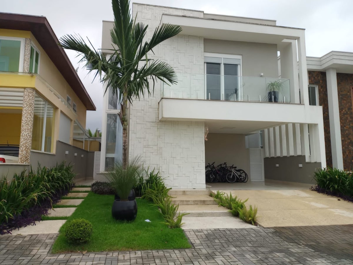 The modern exterior of a two-story house features a clean design with a white facade, large windows, and a balcony. Lush greenery surrounds the entrance, highlighting a well-maintained garden. A paved pathway leads to the front door, providing access to the inviting structure.