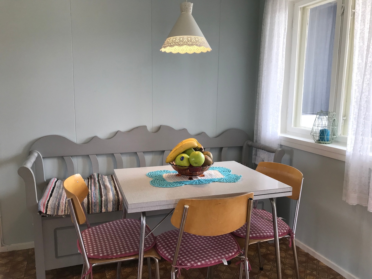 A cozy dining area features a light-colored table with a decorative fruit bowl at the center. Surrounding chairs include four metal-framed options with wooden seats. A gray, cushioned bench against the wall provides additional seating. Natural light filters through a window adorned with white curtains.