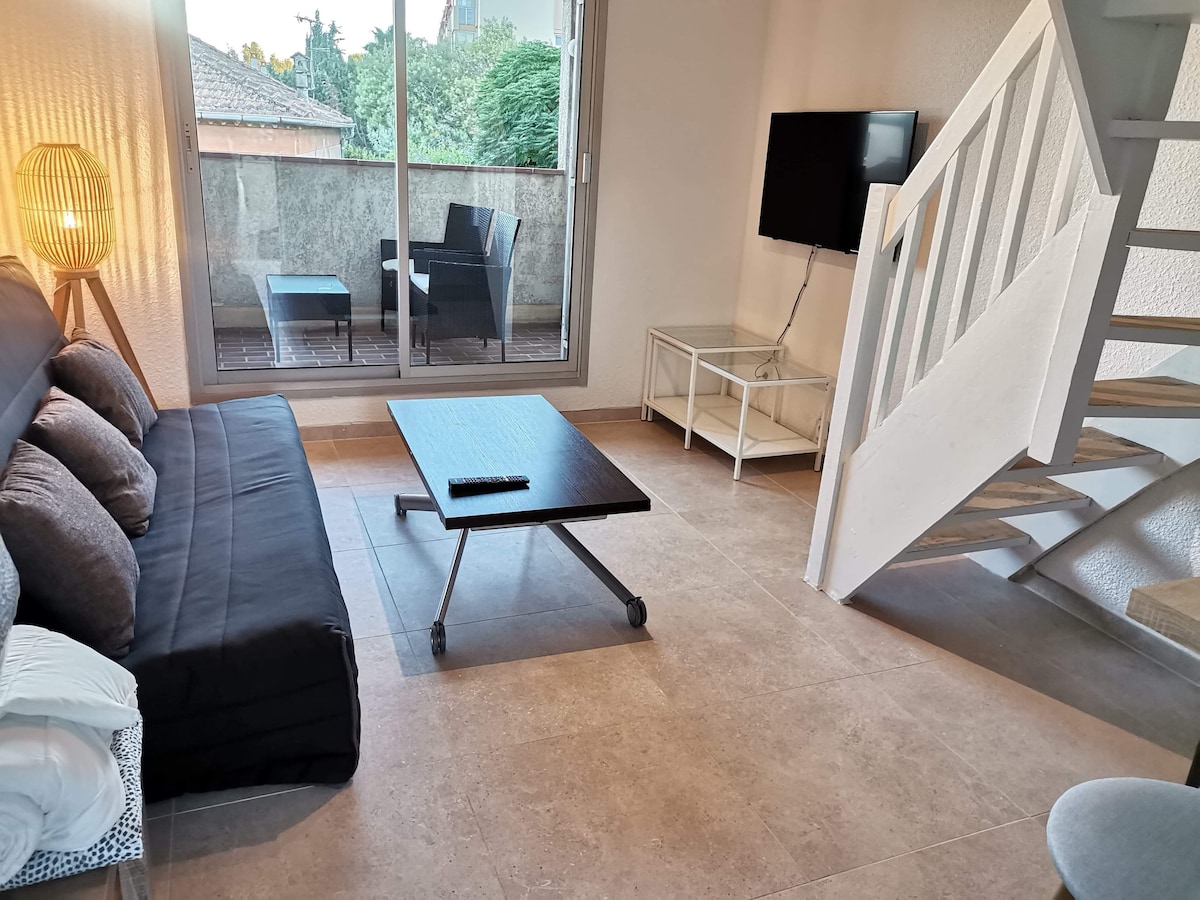 A modern living area features a dark sofa bed accompanied by decorative cushions. A sleek coffee table sits in front, complementing a minimalist white side table. Natural light floods the space through expansive glass doors, revealing a private balcony with seating arrangements. Stairs lead to the upper level.