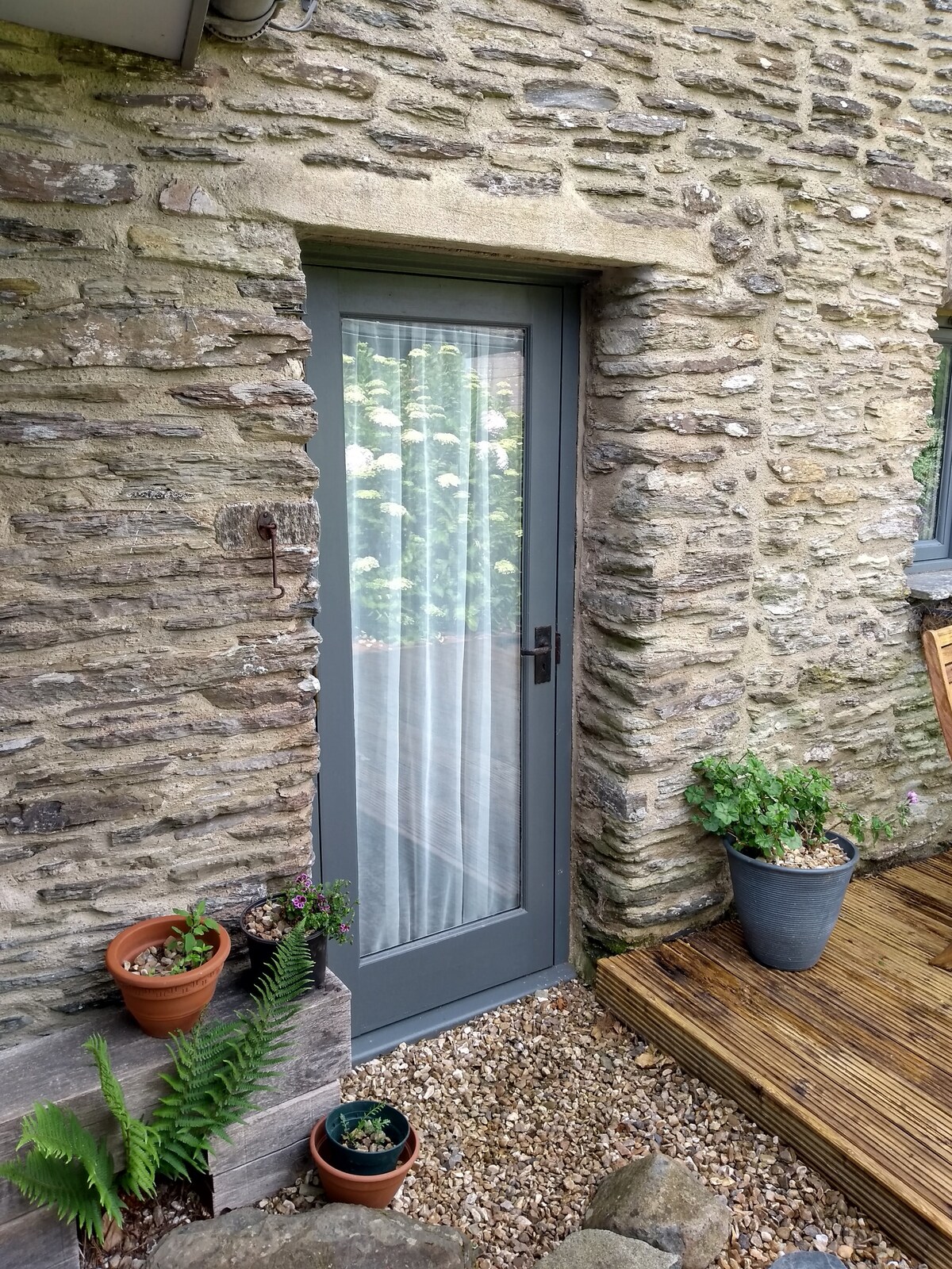 A stone exterior features a grey door framed by sheer curtains, providing a glimpse of the interior. Potted plants and a fern are arranged on either side, while a wooden decking area adds warmth to the entrance. Gravel surrounds the base, enhancing the rustic charm.
