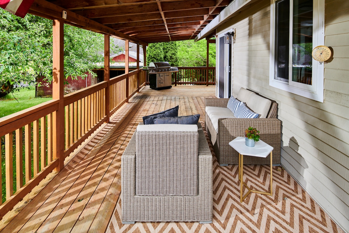 A spacious porch features a patterned flooring and a comfortable seating area with woven chairs and cushions. A small side table holds a plant. In the background, a grill can be seen, surrounded by greenery.