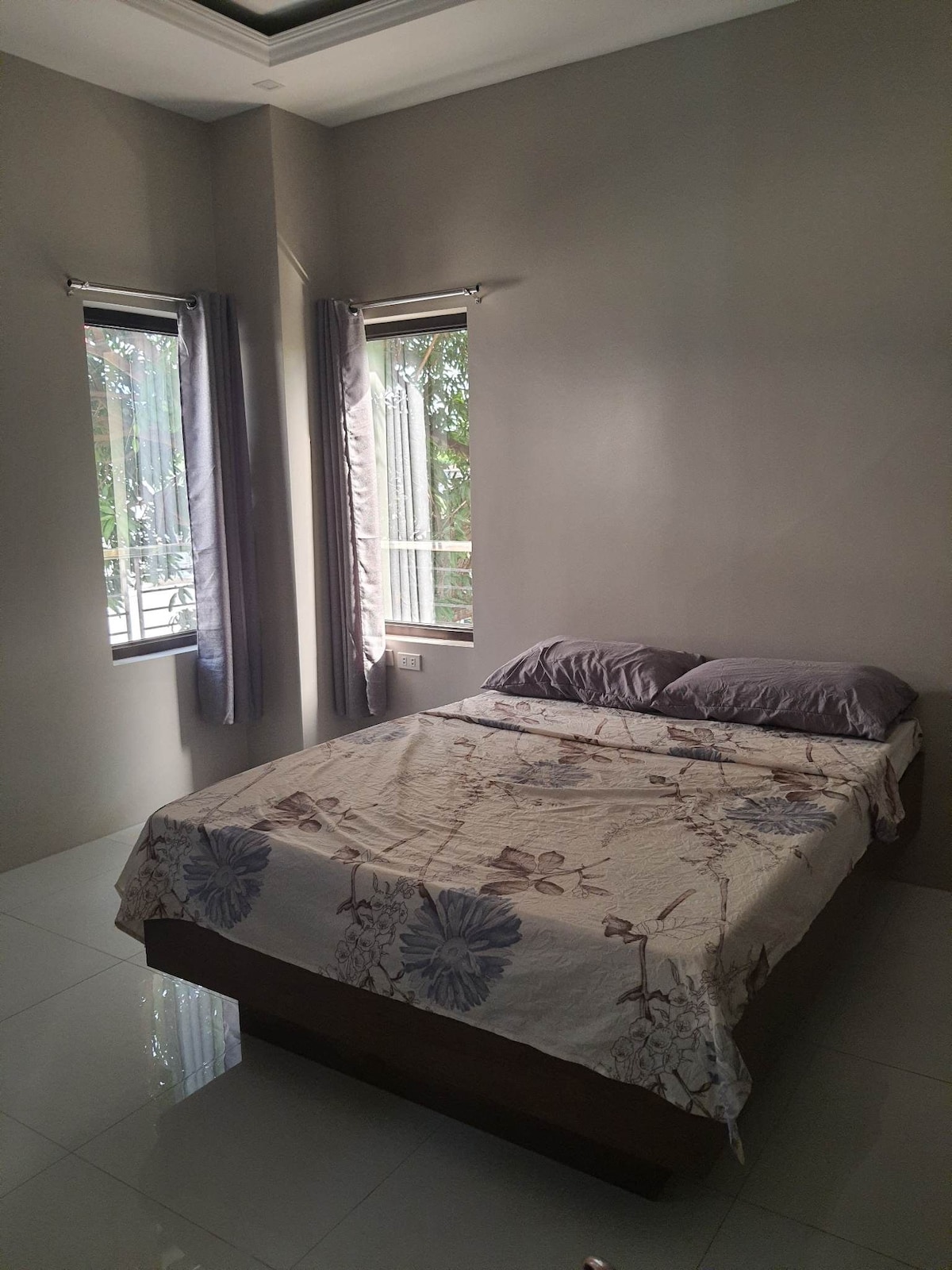 A modest sleeping area features a spacious bed with floral bedding. Natural light enters through two large windows adorned with light curtains, complementing the neutral wall color. The glossy tile floor reflects the room's bright atmosphere.