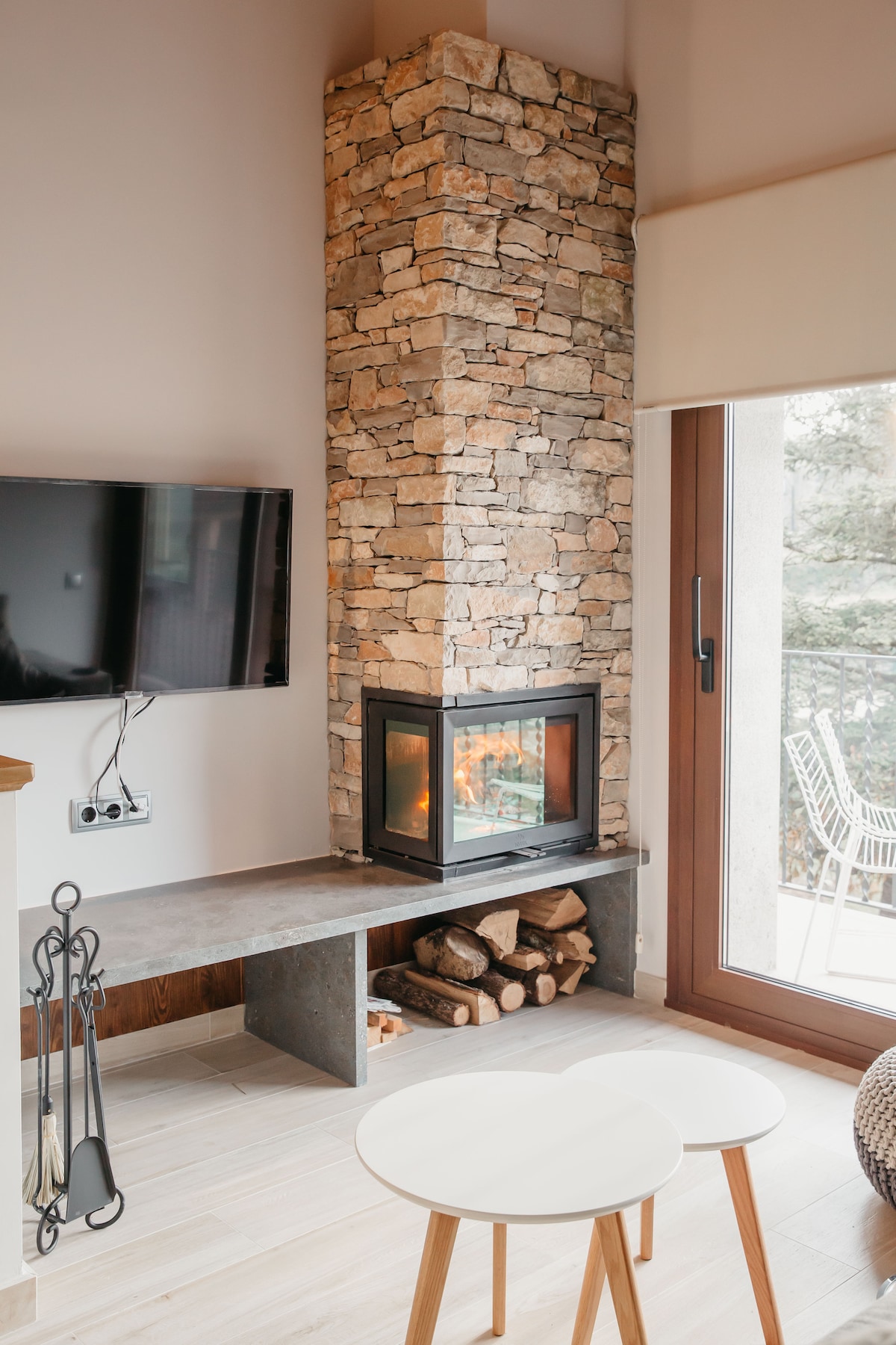 A stone fireplace provides a focal point in the living area, accompanied by a sleek dark shelf and two minimalist white tables. A large window allows natural light to illuminate the space, and logs are neatly stacked beside the fireplace.