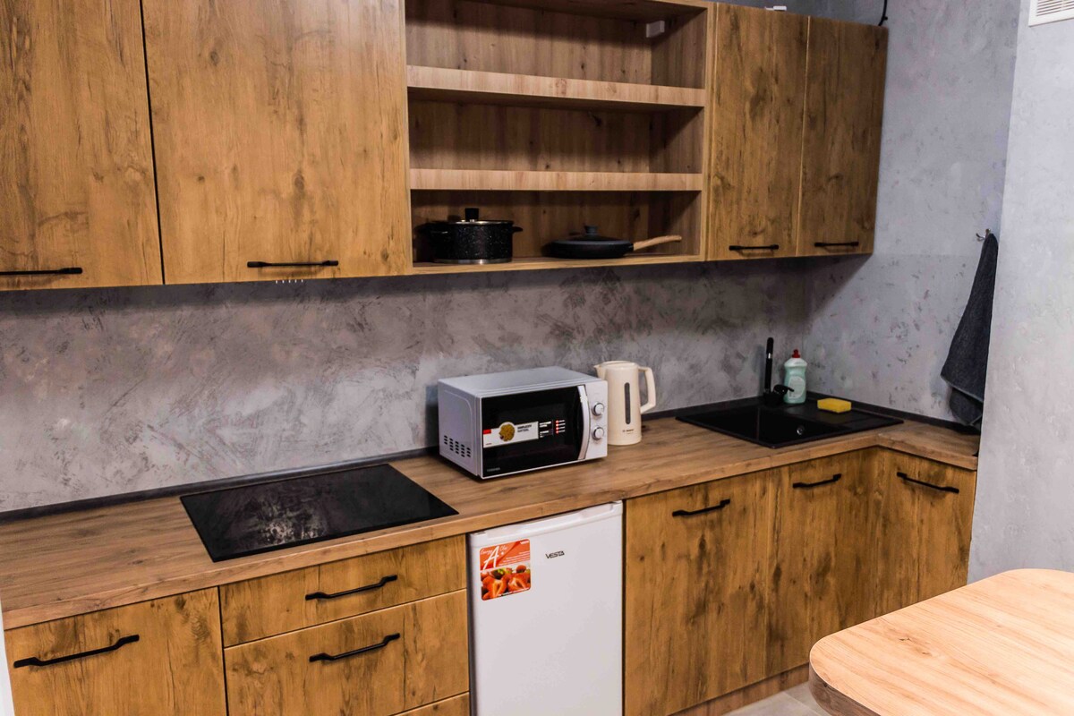 A modern kitchen is presented with warm wood cabinetry and a minimalist design. Essential appliances, including a microwave, kettle, and refrigerator, are neatly arranged. Counter space is available for meal prep, while open shelves provide additional storage options.