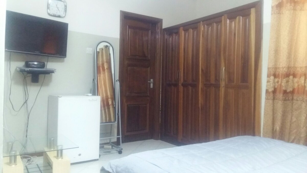 A compact bedroom is presented, featuring a wooden wardrobe with multiple doors alongside a wall-mounted television. A small refrigerator is positioned near a glass table, and a neatly made bed with light bedding occupies the center of the space. Soft curtains frame a window.