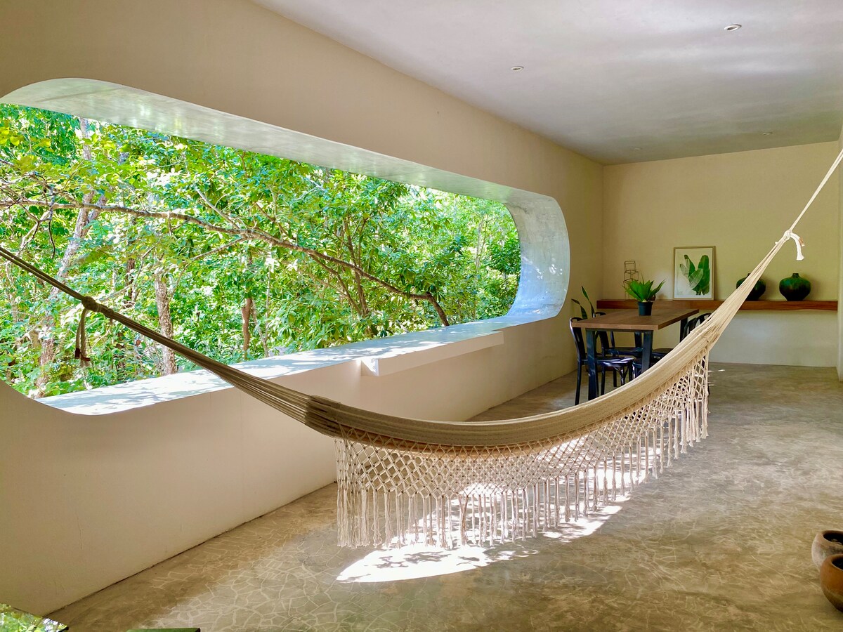 A spacious room features a large window displaying lush greenery from the jungle outside. A hammock is suspended to invite relaxation, while a dining table and chairs are positioned nearby. Natural light illuminates the comfortable space, enhancing the connection with nature.