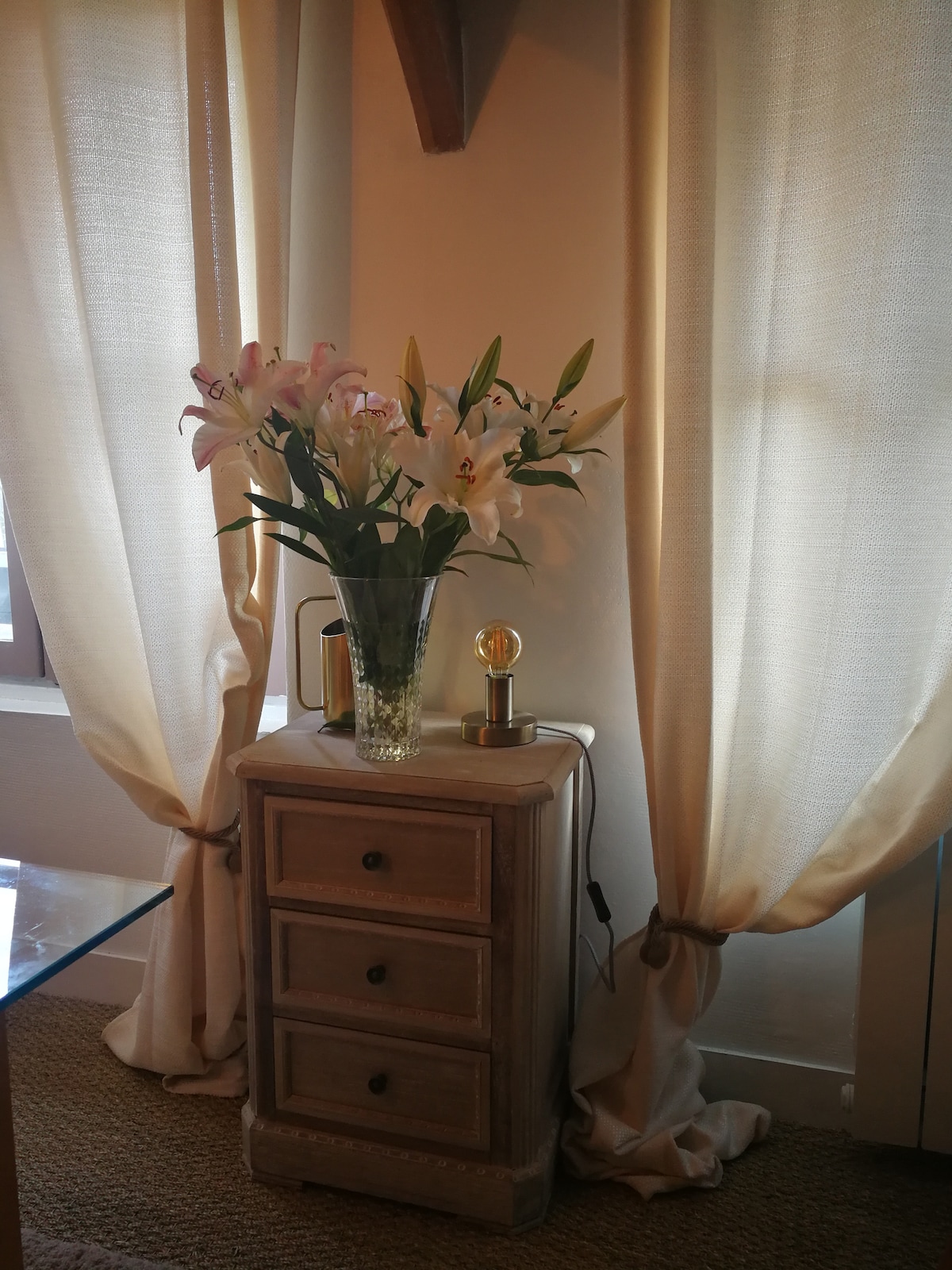 A small wooden side table is positioned next to sheer cream-colored curtains. A glass vase filled with fresh white lilies stands prominently on the table. Soft natural light filters through the window, highlighting the floral arrangement and the textured fabric of the curtains.