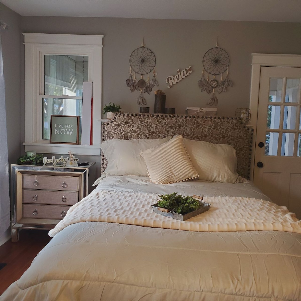 A serene bedroom features a spacious bed dressed in neutral tones with a textured blanket. Decorative dreamcatchers hang above the headboard, and a small tray with greenery rests on the bed. A wooden nightstand with drawers complements the interior's calming aesthetic.