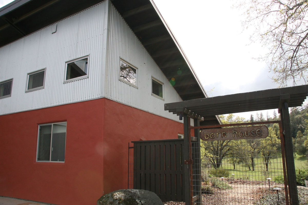 The exterior of the barn house is showcased, featuring a unique design with metal siding and a vibrant red wall. A wooden gate marks the entrance, adorned with a sign labeled 'barn house.' Surrounding greenery and trees create a natural setting.