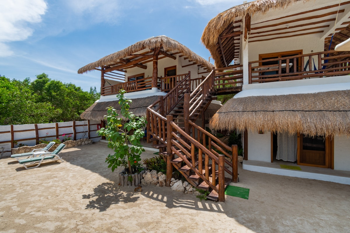 A tropical setting features two-story buildings with wooden balconies and thatched roofs, surrounded by lush greenery. A sandy area includes lounge chairs and a small plant, while a staircase leads to the upper level. Bright blue skies provide a serene backdrop.