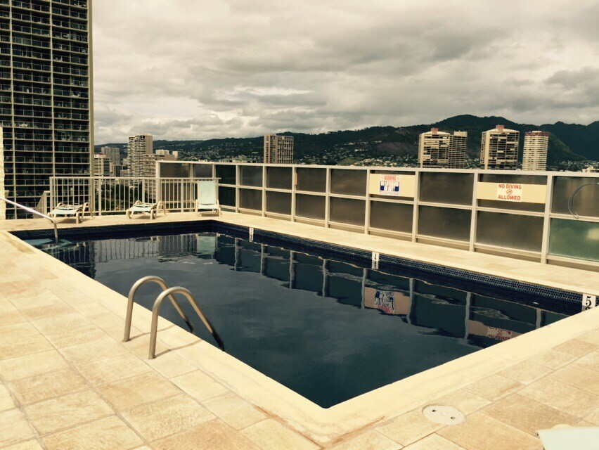 A rooftop pool area is presented, featuring a rectangular swimming pool with steps for entry. Surrounding the pool, lounge chairs are arranged for relaxation. The backdrop includes city views and distant mountains, creating a serene ambiance.