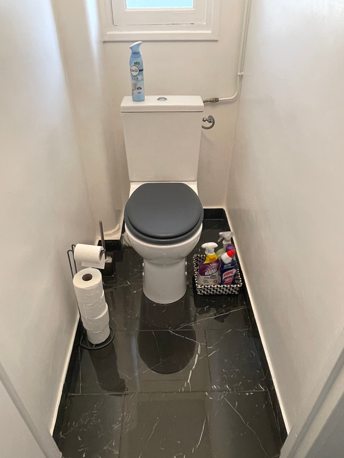 A compact bathroom space features a modern toilet with a comfortable seat. Cleaning products are neatly arranged on the floor along with a basket containing toiletries. White walls and dark tiled flooring create a clean, bright atmosphere.