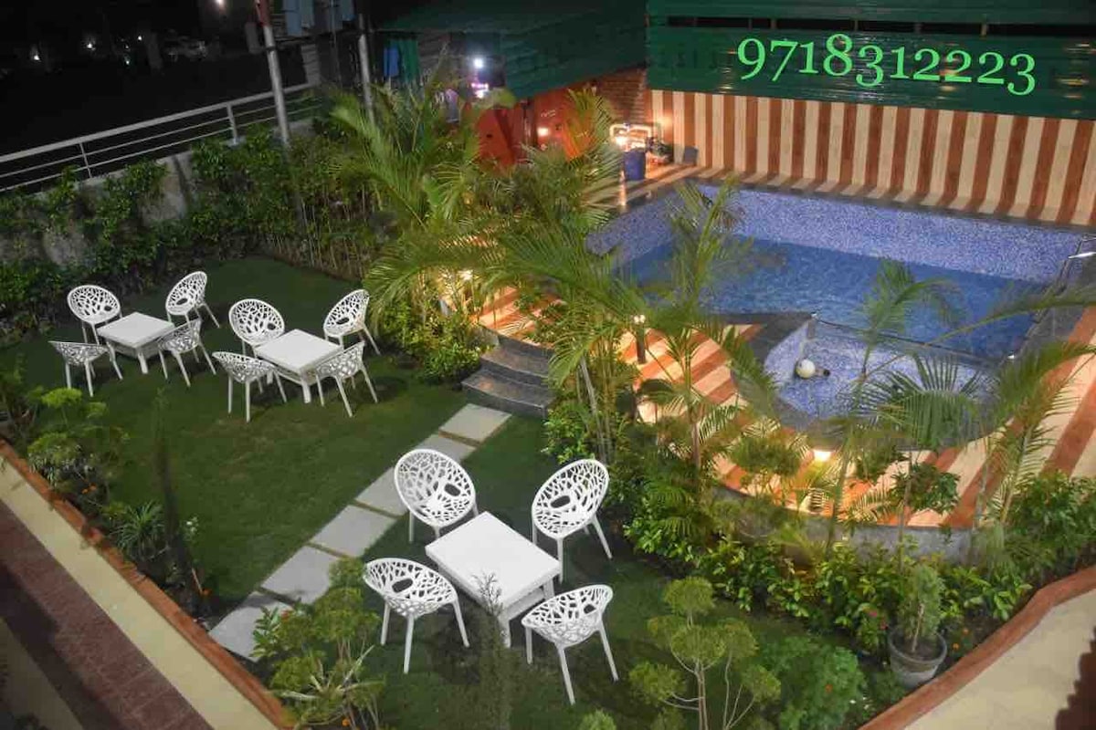 An inviting pool area is surrounded by lush greenery and vibrant plants. Several white outdoor tables and chairs are arranged on a well-maintained lawn, providing comfortable seating options. Soft lighting enhances the tranquil atmosphere, creating a welcoming space for relaxation.