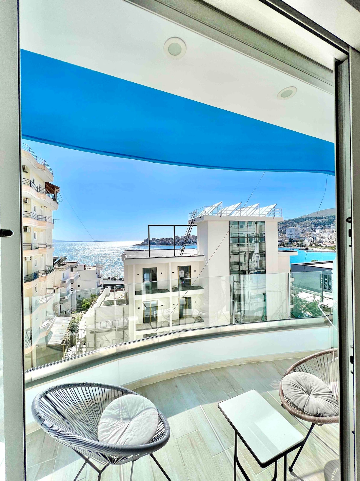 A balcony offers a panoramic view of the sea, with glass railings ensuring an unobstructed vista. Two modern chairs are positioned beside a small table, inviting relaxation. Bright blue awnings provide shade, contrasting with the white architecture of surrounding buildings and the clear sky.