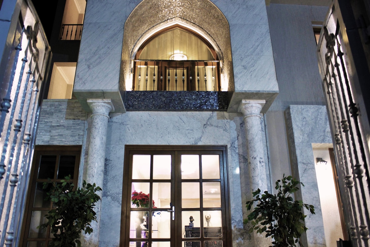 The villa's entrance is framed by elegant marble columns, showcasing intricate architectural details. A large arched window is situated above a set of double doors, with warm lighting illuminating the façade. Potted plants add a touch of greenery to the welcoming entrance.