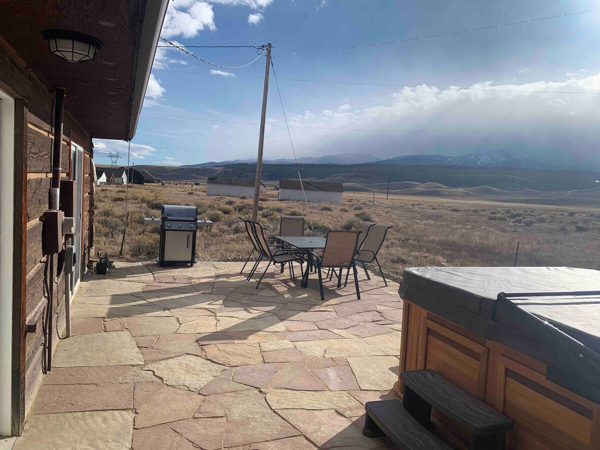An outdoor patio area is visible, featuring a stone floor and a table with several chairs. A grill is positioned nearby. The expansive landscape includes mountains and open fields under a partly cloudy sky, creating a serene environment.