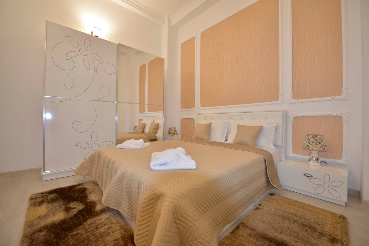A spacious room features a plush double bed dressed in neutral linens, accompanied by decorative pillows. A mirrored wardrobe enhances the sense of openness. Soft, warm hues adorn the walls, while a small lamp and a cozy area rug add a touch of comfort.