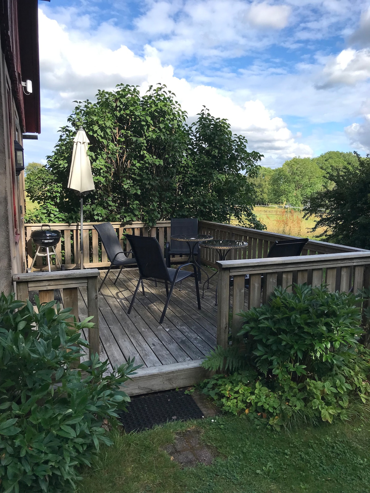An outdoor deck is positioned amidst greenery, featuring a wooden surface. Four black chairs surround a small table, while a grill is located nearby. A large umbrella provides shade, and the setting is complemented by lush bushes and trees in the background.