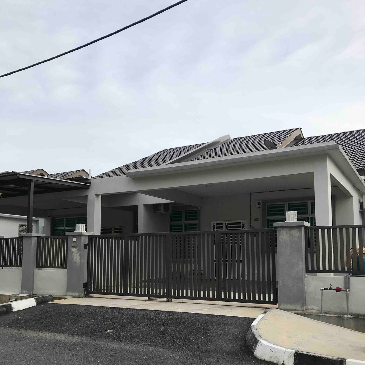 The exterior of a single storey semi-detached house is displayed, featuring a sloped roof and a modern grey facade. The property is enclosed by a horizontal metal gate, with a driveway and surrounding pavement visible. A cloudy sky is seen overhead.