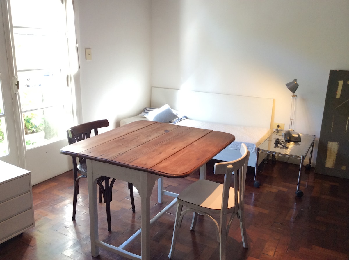 A simple living room features a wooden table accompanied by two chairs, one wooden and one metal. A spacious bed is situated against the wall, with a desk lamp and a typewriter on a small mobile cart nearby. Natural light enters through glass doors.