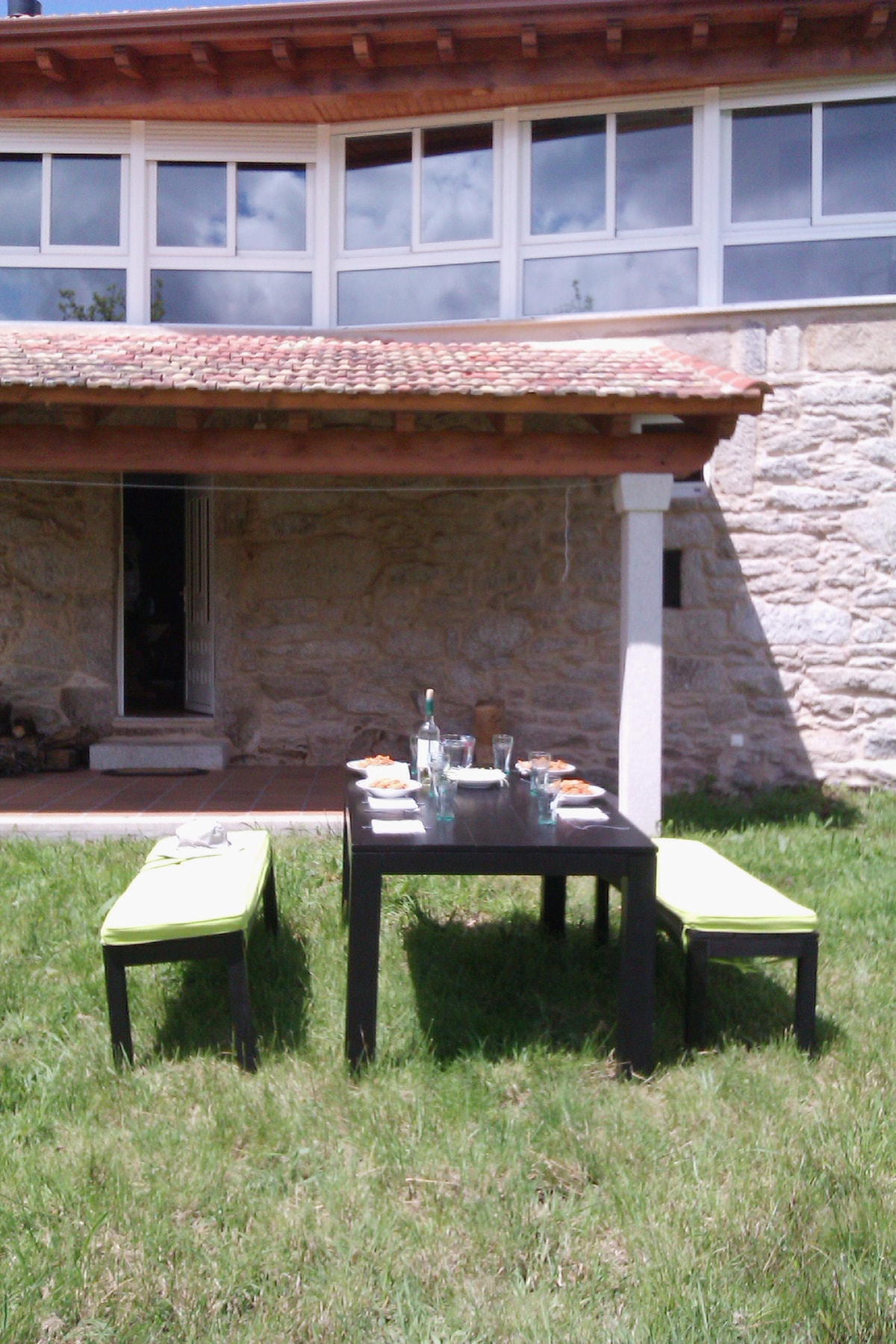 A dining table is set on a grassy area beneath a covered porch. Two green benches flank the black table, which holds dishes and drinks. The stone exterior of the house and large windows provide a rustic yet modern backdrop.